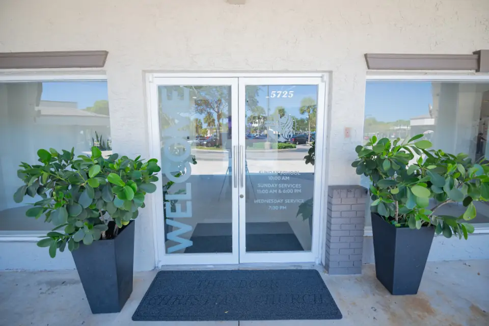 The glass entrance doors of Sacred Farewells at 5725 South University Drive with planters on either side